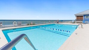 Una piscina cristallina blu circondata da una recinzione metallica di sicurezza, con una postazione di salvataggio e un edificio con tetto blu sullo sfondo.