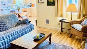 A living room with a sofa, a coffee table, and a wicker chair. There is a kitchen in the background with wooden cabinets and a dining table. - Peaceful, Fully Equipped Cabin Near Beach & Town, Certified NWF Wildlife Habitat (Seaview)