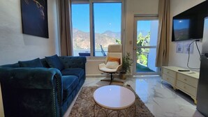 Uma sala de estar com um sofá azul, uma poltrona branca, uma mesa redonda pequena e uma TV fixada na parede.