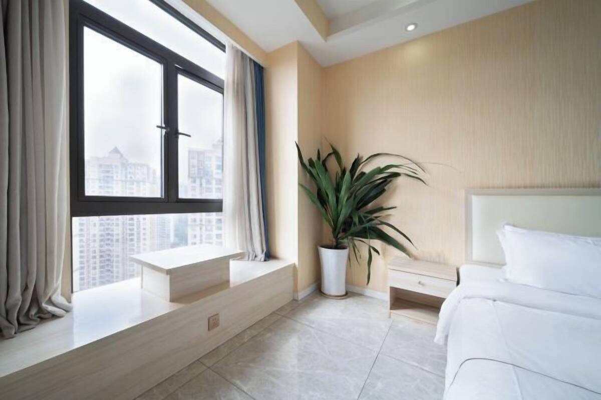 Ein modernes Hotelzimmer mit großem Fenster, einer Zimmerpflanze, einem Bett und einem kleinen Nachttisch.
