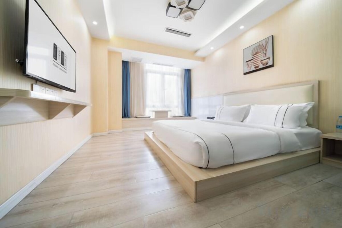 A modern hotel room with a large bed, a flat-screen TV mounted on the wall, and a window with curtains.