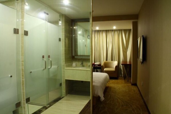 A hotel room with a glass-enclosed shower, a bed, a chair, a desk, and a television.