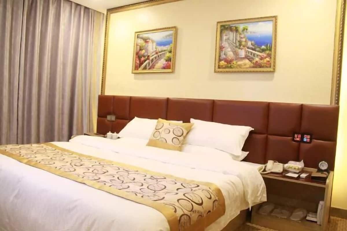 A hotel room with a large bed, a headboard, two paintings on the wall, a nightstand, and a curtain.
