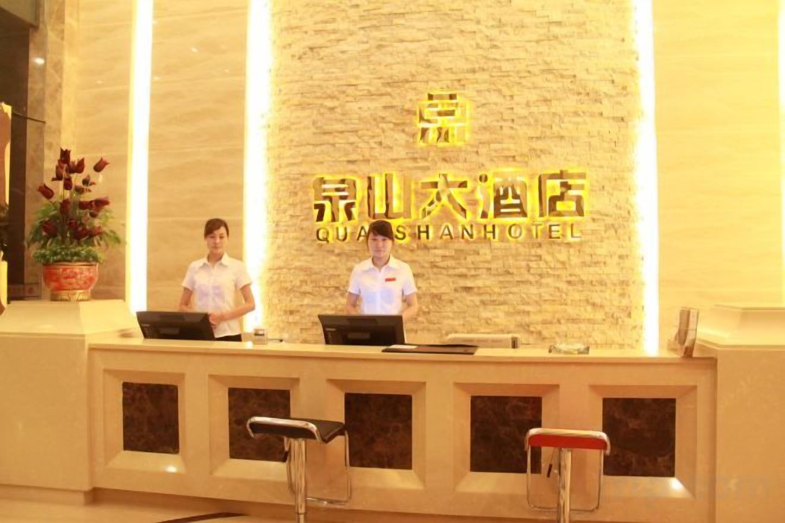 A hotel reception desk with two staff members, a stone wall with hotel signage, and a potted plant.