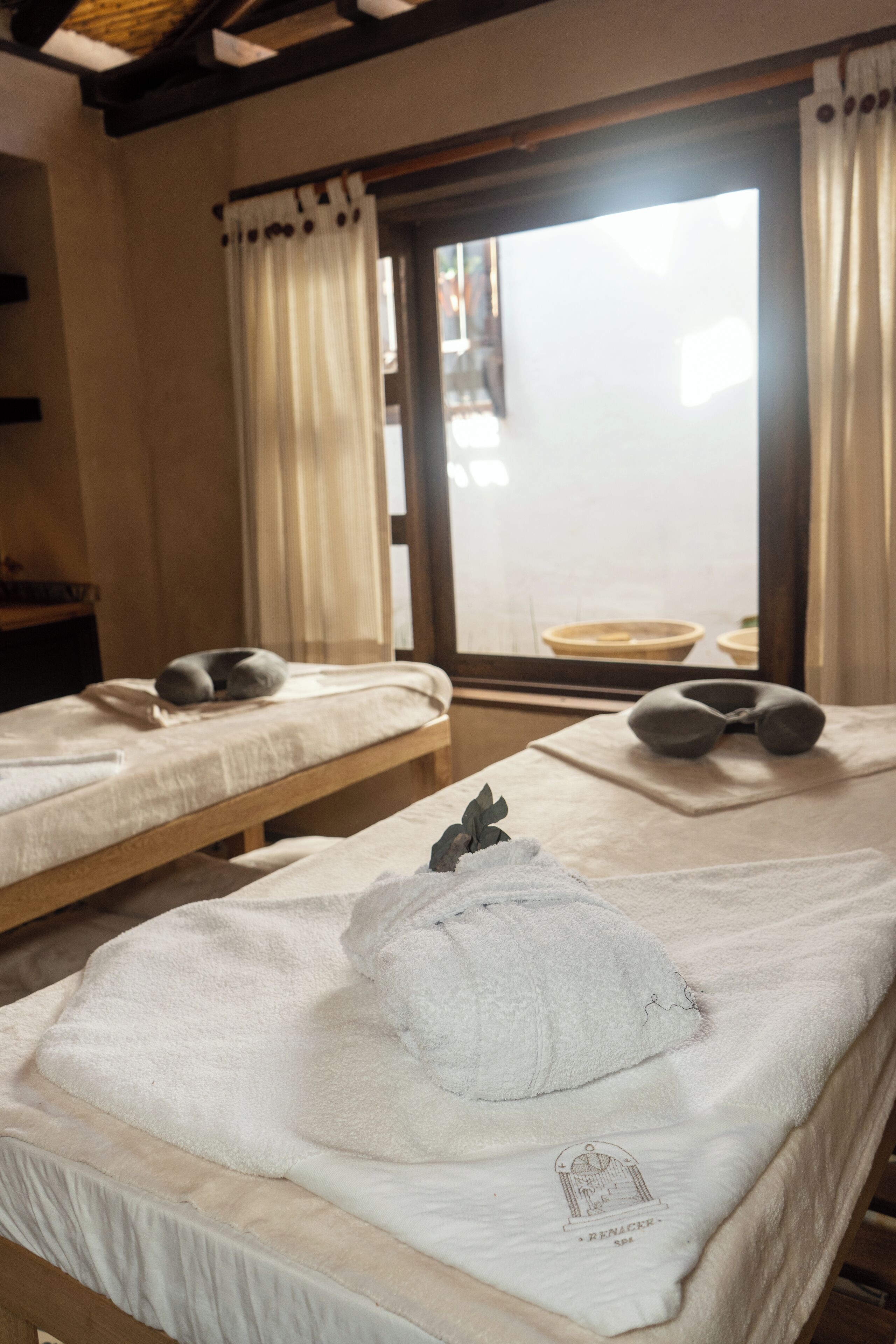 Una habitación con dos camas, cada una con una almohada y una toalla enrollada. Hay una ventana con cortinas y vistas al exterior.