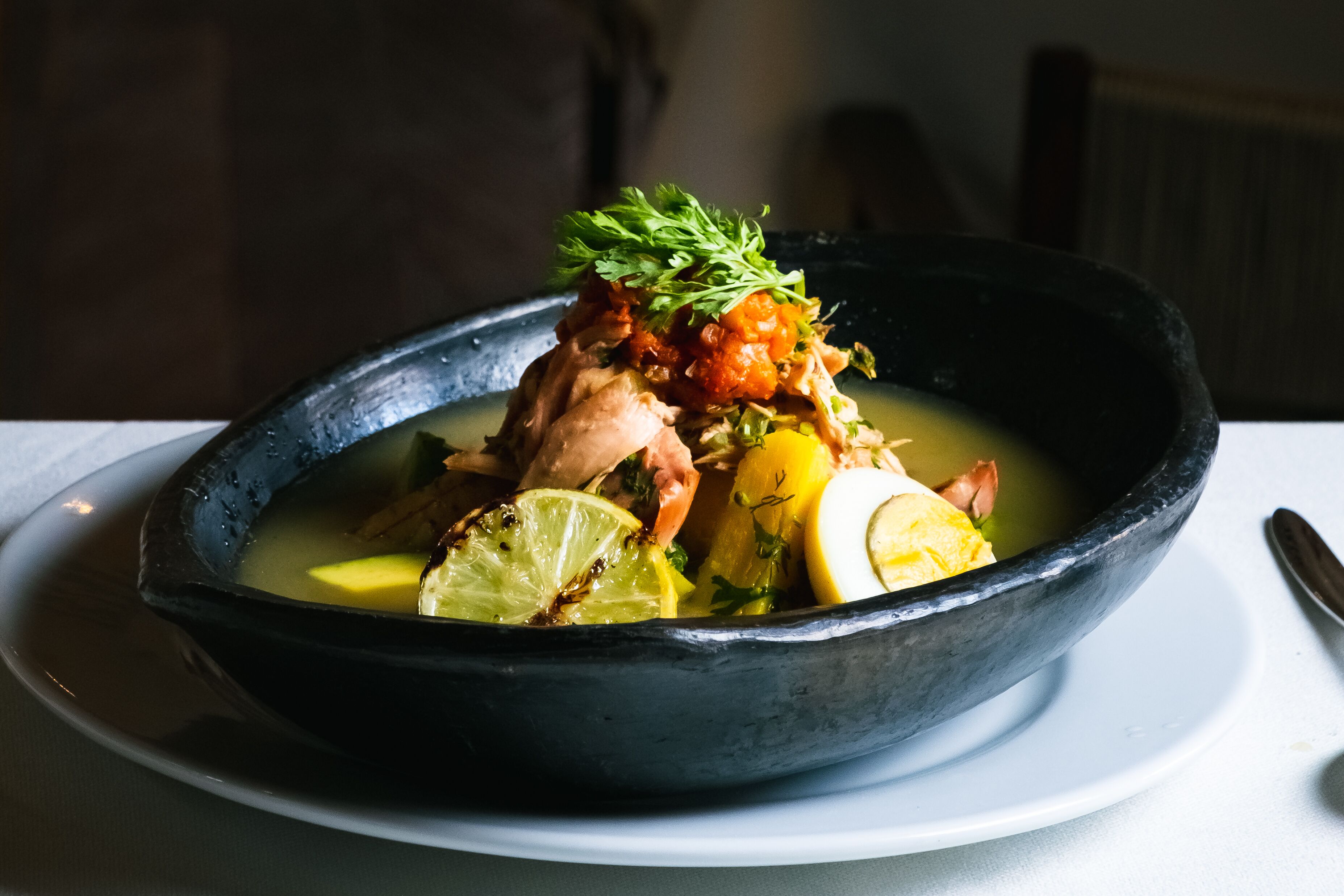 Un tazón de sopa con diversos ingredientes, incluyendo un trozo de carne, limón y hierbas, servido en un recipiente oscuro sobre un plato blanco.