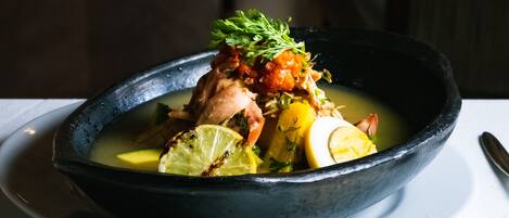 Un tazón de sopa con diversos ingredientes, incluyendo un trozo de carne, limón y hierbas, servido en un recipiente oscuro sobre un plato blanco.
