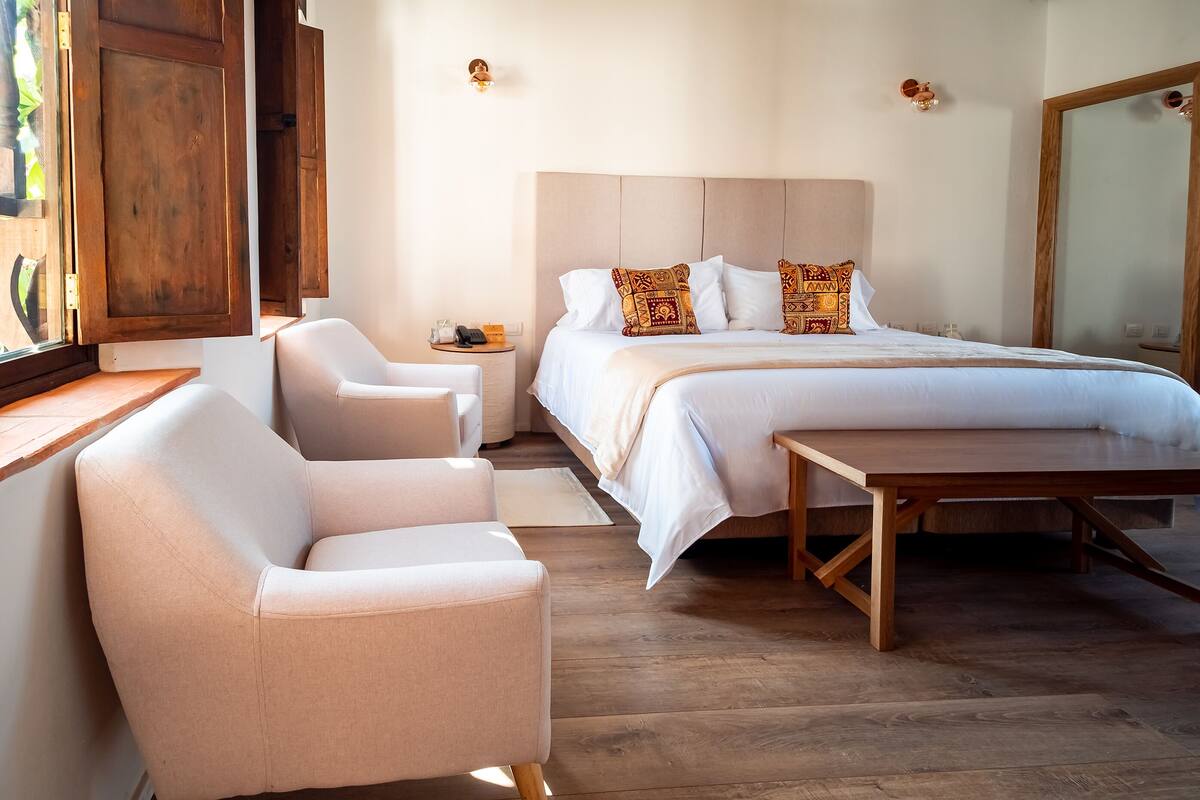 A bedroom with a bed, two chairs, a wooden table, a nightstand, and a mirror.