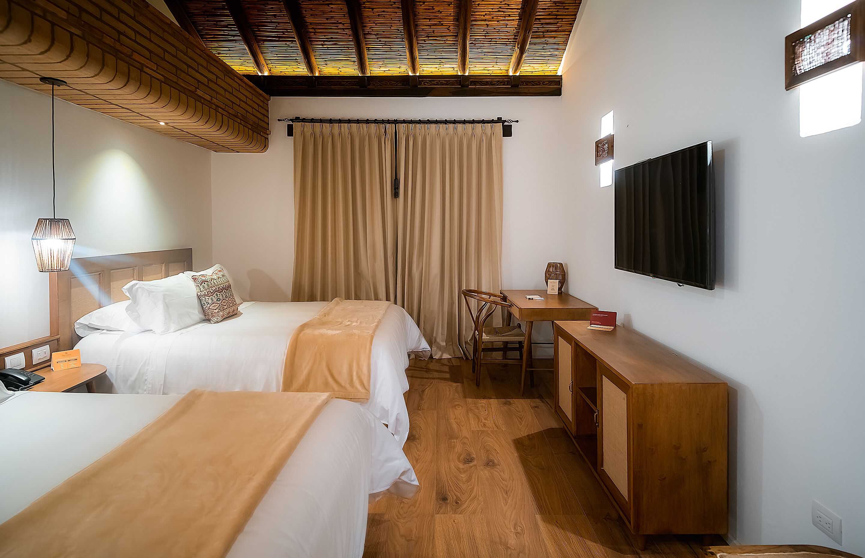 A hotel room with two beds, a desk, a chair, a TV, and a wooden ceiling.