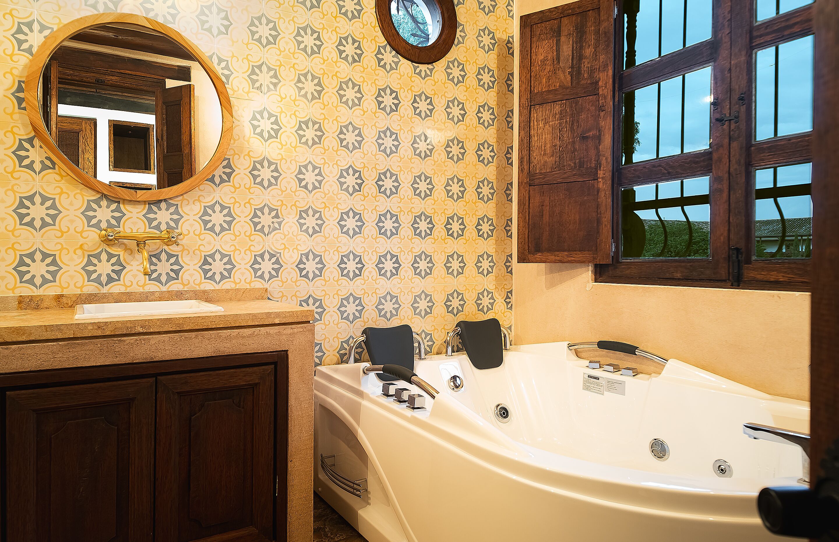 Un baño con una bañera grande, armarios de madera, un espejo redondo y papel tapiz estampado.