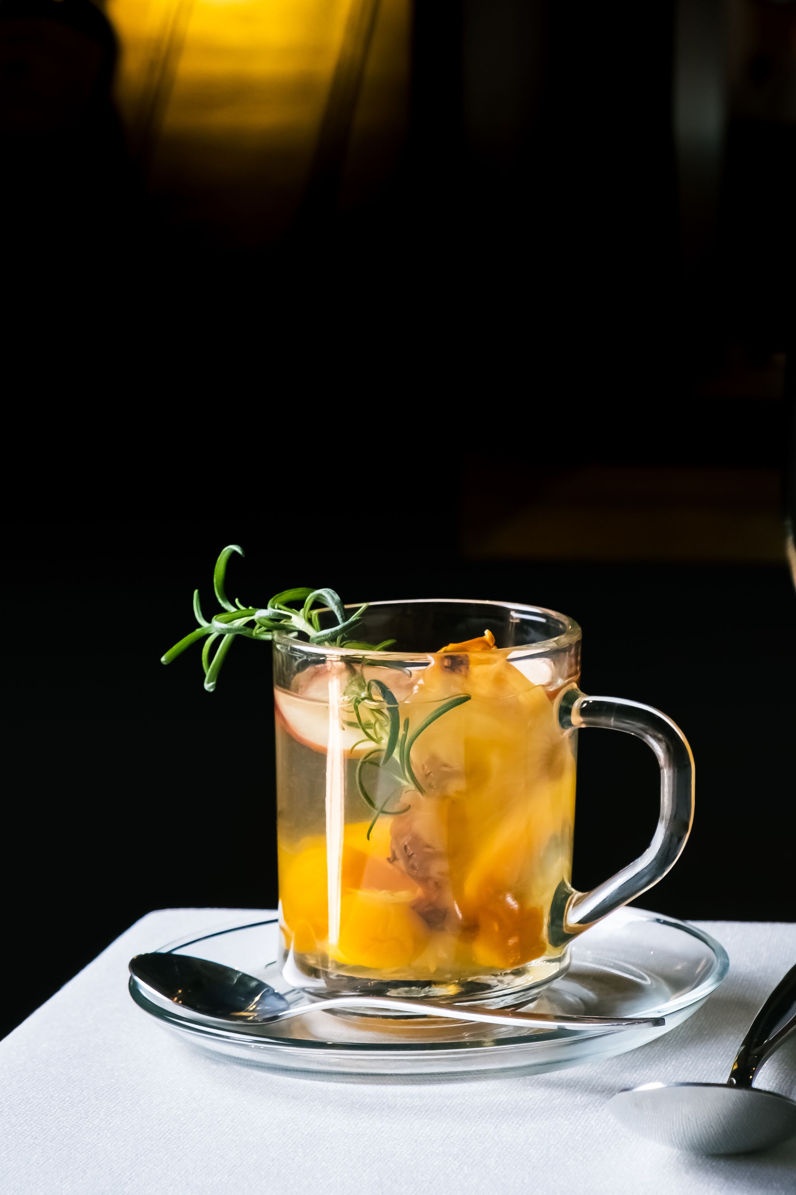 A clear glass mug with a handle, filled with a yellowish liquid, possibly tea, and garnished with a sprig of rosemary.