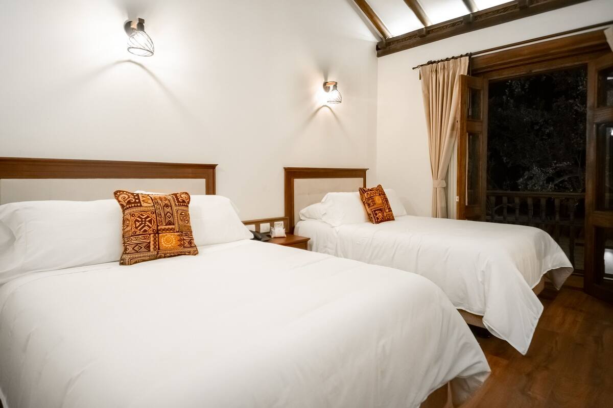 A room with two beds, each with white bedding and patterned pillows. There are wall-mounted lights and a window with curtains.