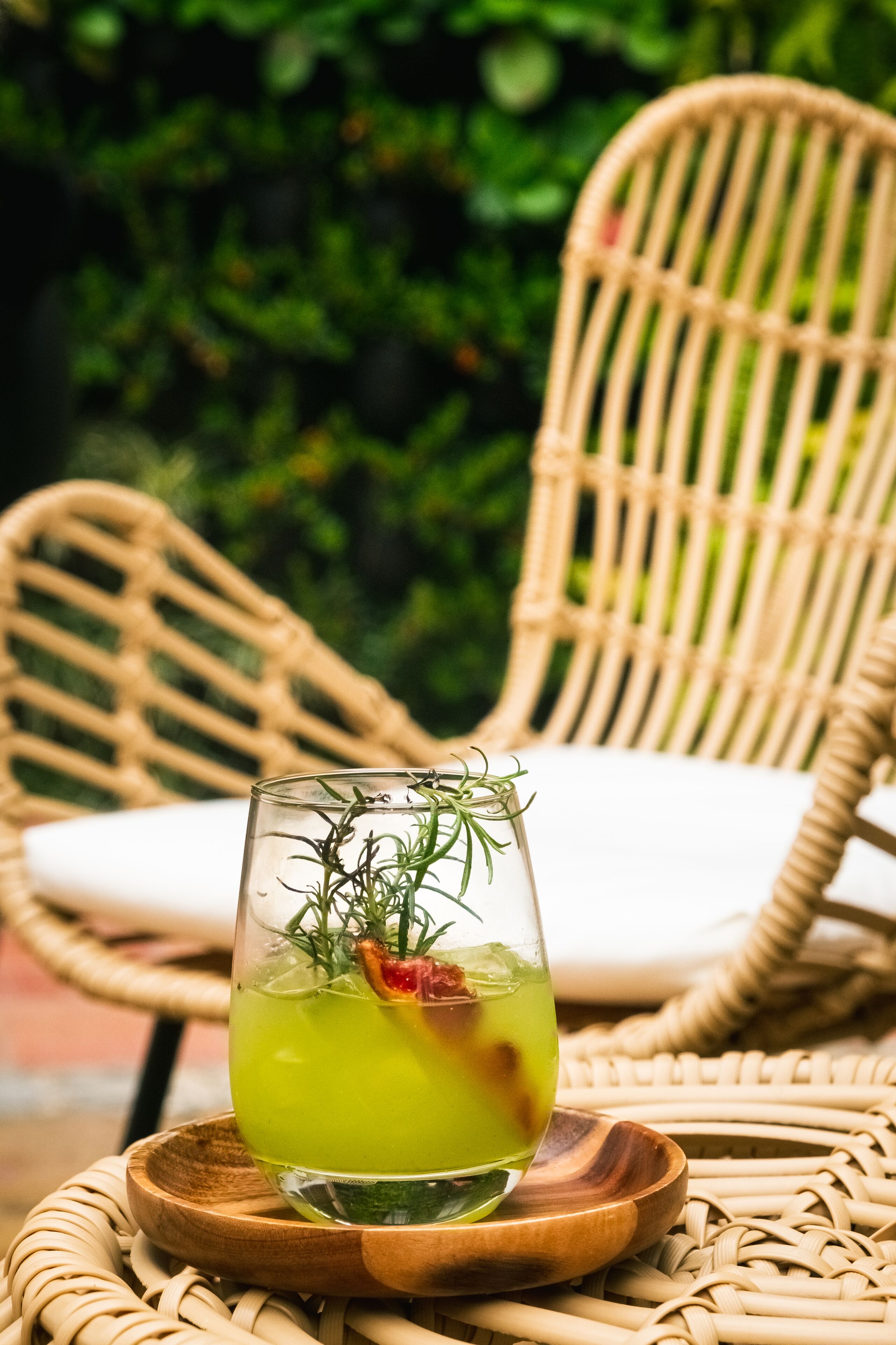 Une chaise en osier avec un coussin blanc, un verre d’une boisson verte agrémentée d’une garniture, et un sous-verre en bois.