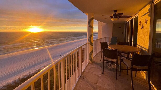 Un balcon avec vue sur le coucher de soleil sur l’océan, une table avec des chaises et un ventilateur de plafond.