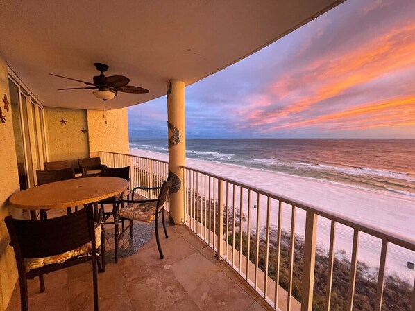 Ein Balkon mit Tisch und Stühlen, der Blick auf einen Strand mit Sonnenuntergang bietet.