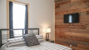 A bedroom with a bed, a nightstand, a lamp, a TV mounted on a wooden wall, and a window with curtains. - Private, quiet & beautiful second story with peek a boo water views (Milbridge)
