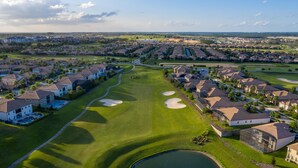 Vue aérienne d’un terrain de golf avec des immeubles résidentiels, un étang et un ciel dégagé.