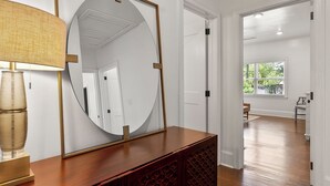 A room with a large mirror, a wooden console table, and a lamp. There is a door leading to another room with a window and a view of greenery outside.