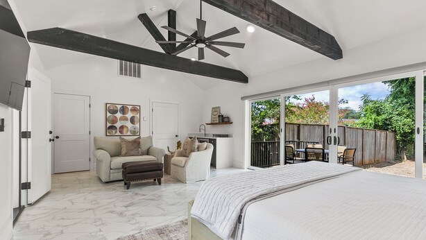 A spacious bedroom with a large bed, a ceiling fan, a seating area with a sofa and chairs, and a view of the outdoors through large sliding glass doors.