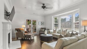 A spacious living room with a fireplace, a ceiling fan, and a large window with blinds.