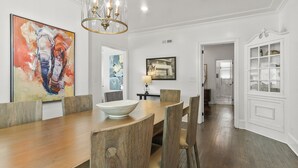 A dining area with a wooden table, chairs, and a chandelier. There are paintings on the walls and a door with glass panels.