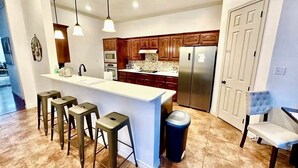 A modern kitchen with a central island, bar stools, a trash can, and a dining area with a chair. - 10th St Manor + Walk to Stadium + Hot Tub + Game Garage + Sleeps 20 (Lubbock)
