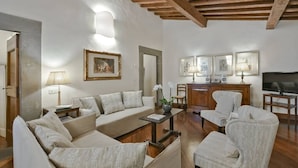 A living room with a sofa, armchairs, a coffee table, and a TV. There are framed pictures on the wall and a lamp on the side table.