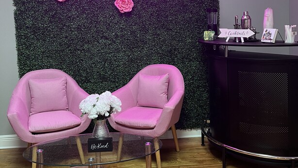 A room with a neon sign reading "Lady B's Place," a green wall with pink flowers, two pink chairs, a small table with a vase, and a black cabinet with items on top.