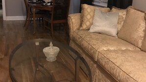 A living room with a glass coffee table, a beige sofa, a dining table with chairs, and a chandelier.