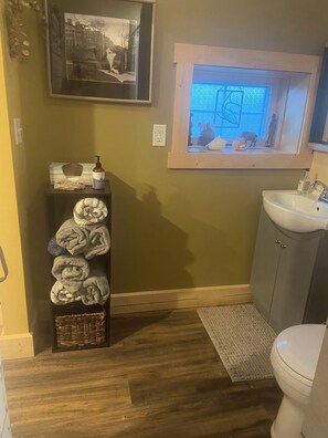 A bathroom with a wooden floor, a sink, a toilet, a window with a view, a shelf with towels, and a framed picture on the wall.