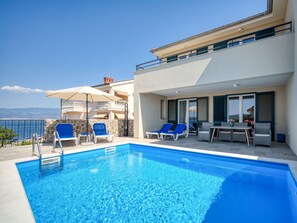 Ein Swimmingpool mit Liegestühlen und Sonnenschirmen, ein Esstisch und Blick aufs Meer.