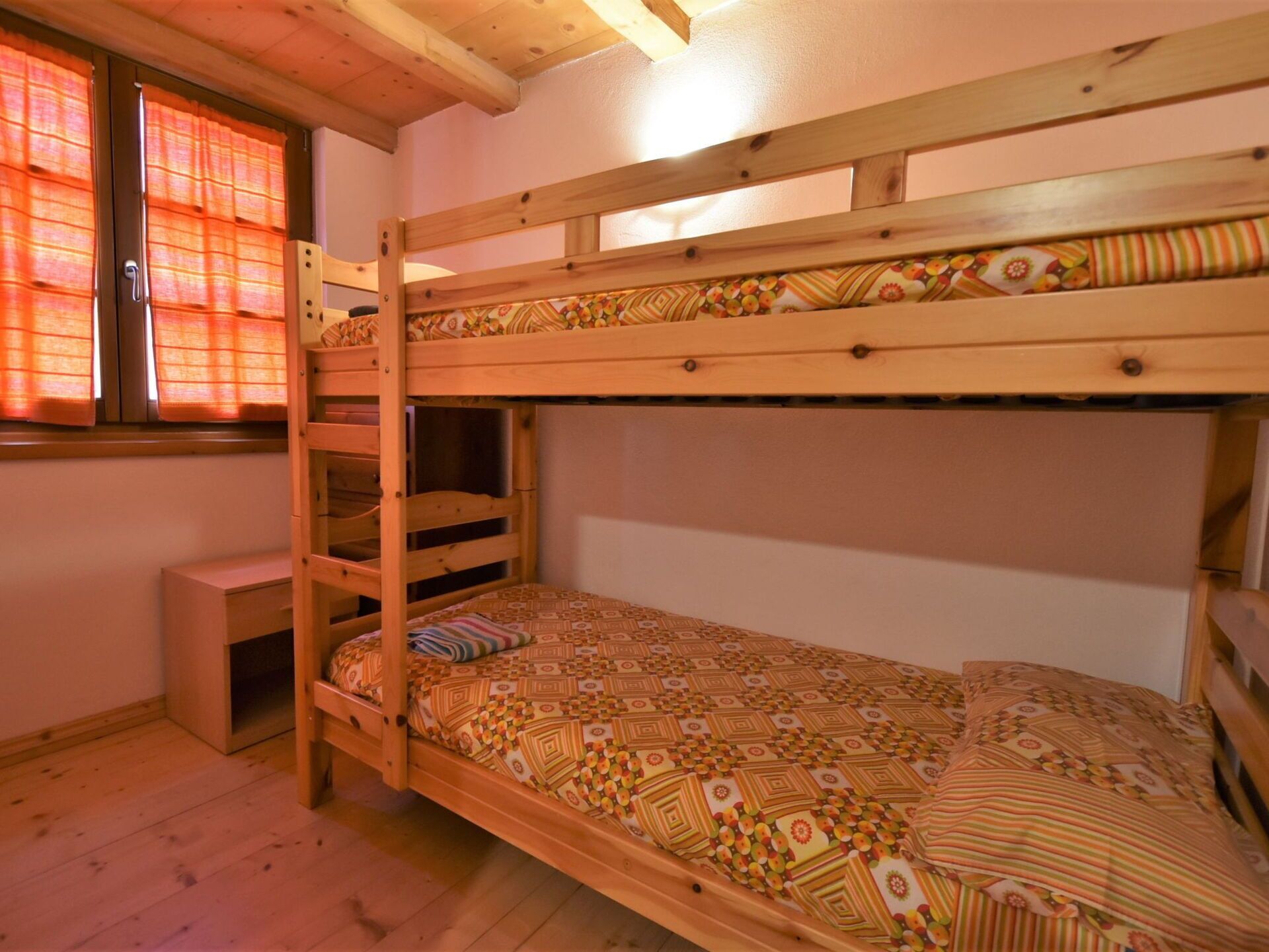 Habitación con literas de madera, una mesita de noche pequeña y una ventana con cortinas anaranjadas.