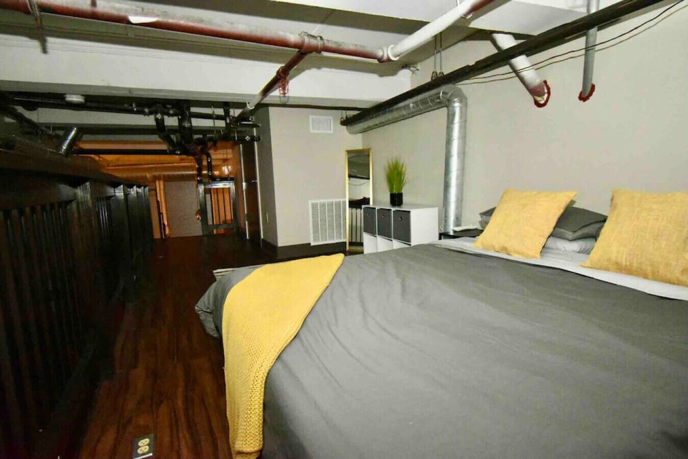 A room with a bed, a wooden floor, a radiator, and visible pipes on the ceiling.