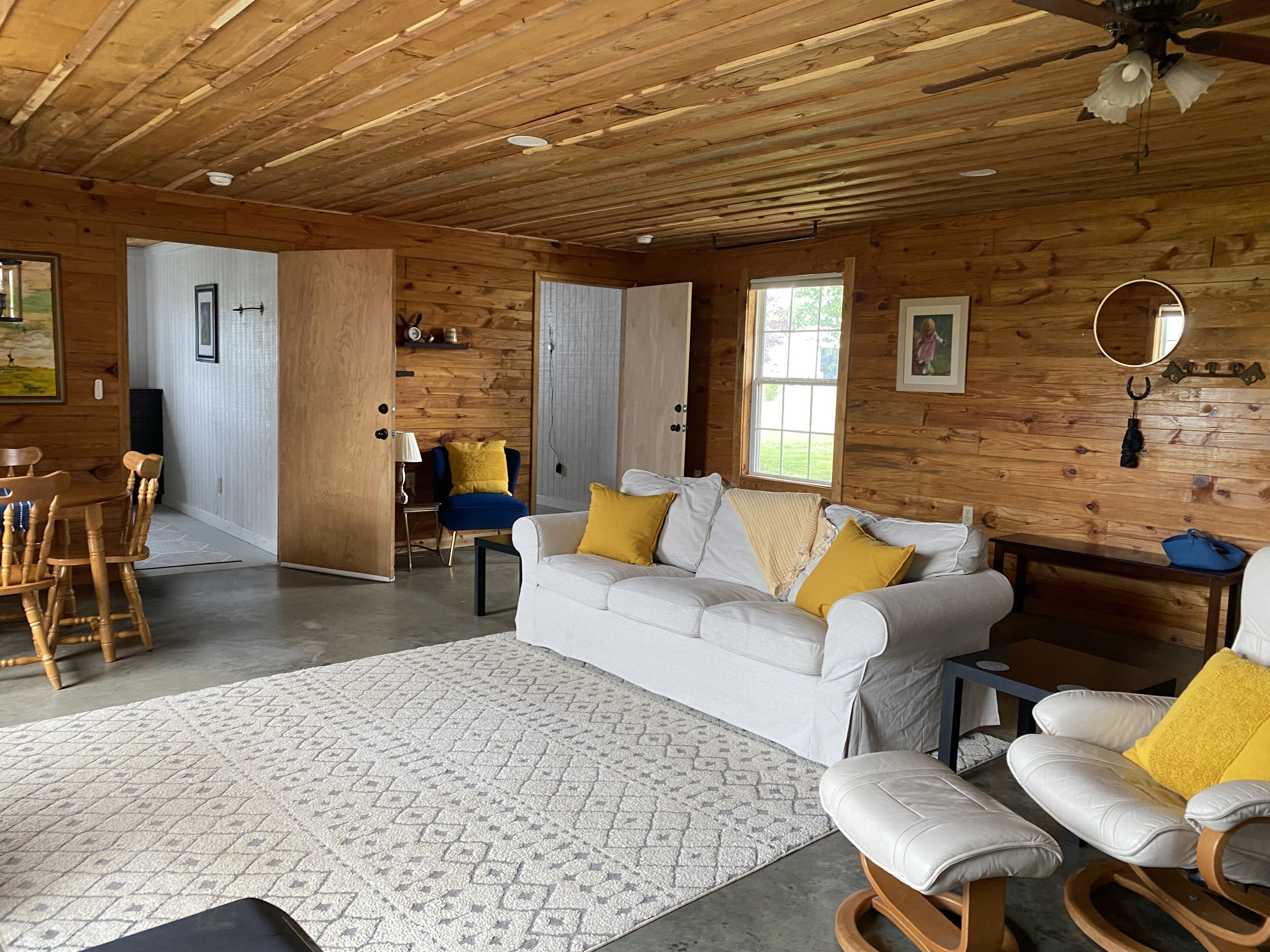 A wooden cabin interior with a white sofa, yellow cushions, a rocking chair, a dining table with chairs, and a patterned rug.