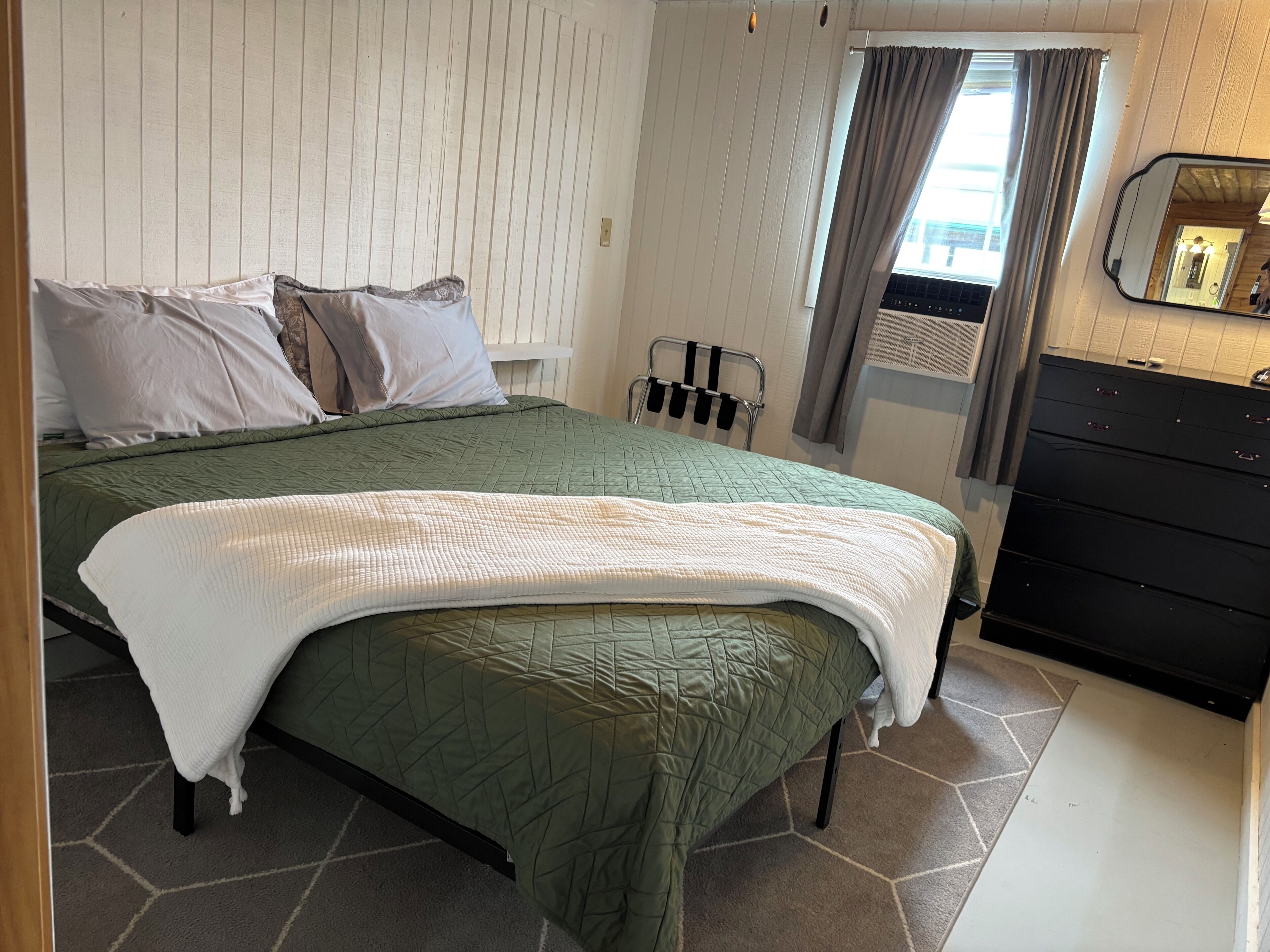 Une chambre à coucher avec une couette matelassée verte, des oreillers blancs, un couvre-lit blanc, une tête de lit en métal, une fenêtre avec des rideaux, un miroir et une commode noire.