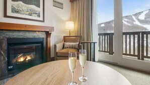 Ein gemütliches Zimmer mit Kamin, einem runden Holztisch mit zwei Sektgläsern, einer bequemen Couch und einem Balkon mit Bergblick.