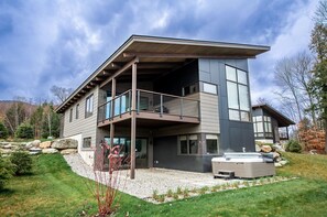 A modern two-story house with a balcony, large windows, and a hot tub in the front yard. - Modern Mountain Home at Wildewood (Stowe)