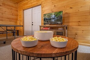 Interior de uma cabana de madeira com uma mesa redonda de madeira, três tigelas brancas de pipoca, uma televisão sobre um suporte de madeira e uma porta branca.
