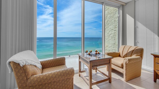 Una habitación con un gran ventanal con vistas al océano, dos sillones, una pequeña mesa con velas y suelo de madera.