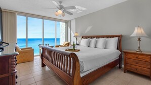 Habitación de hotel con una cama grande, una mesita de noche de madera, un escritorio y una silla. Se ve el océano a través de ventanas amplias.