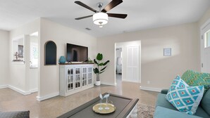A spacious living room with a ceiling fan, a television, a coffee table with decorative items, a sofa with patterned cushions, and a potted plant. - Here Comes The Sun! King Bed - One block to the water Unit #4 (St. Augustine)