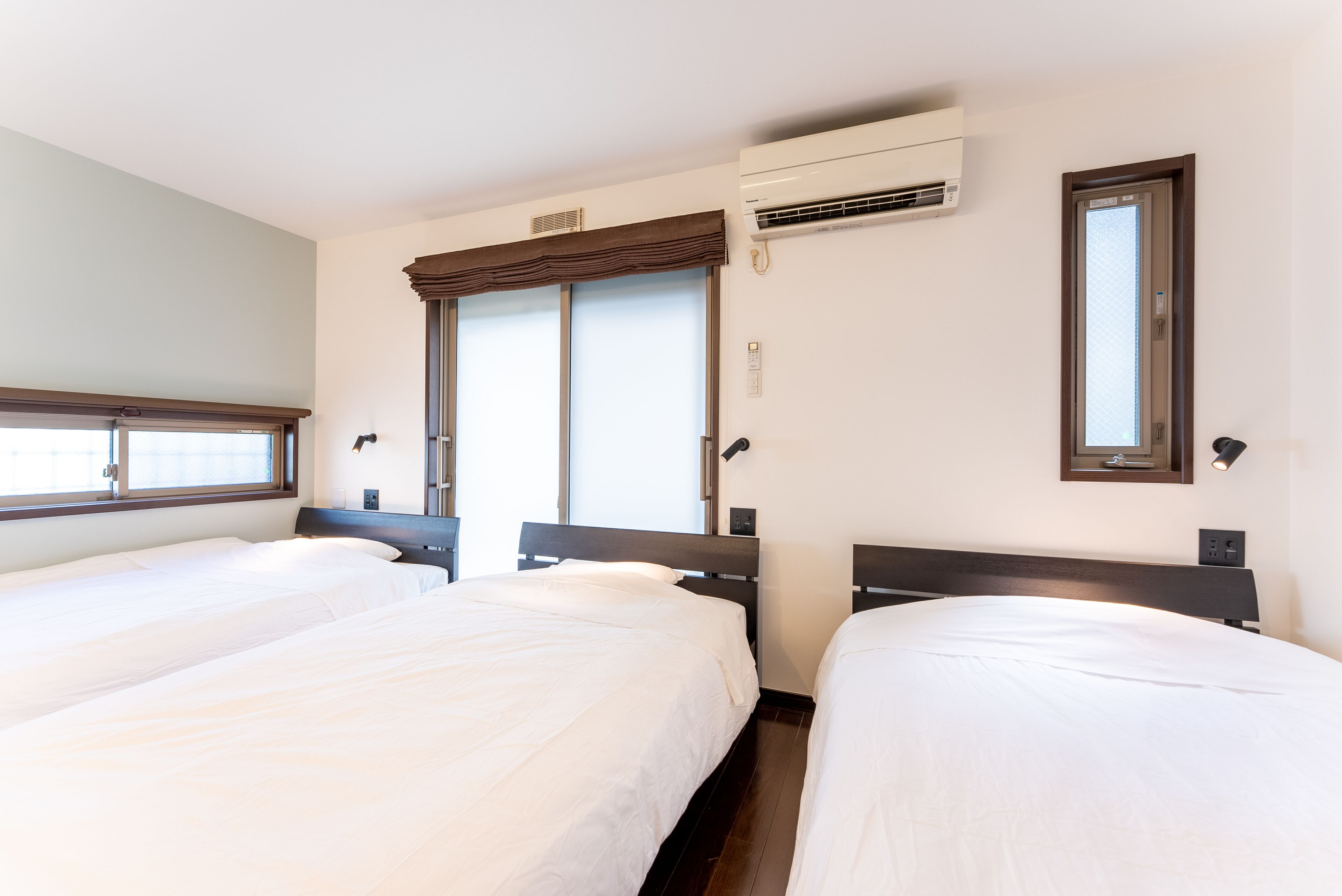 A hotel room with two beds, a large window with blinds, an air conditioning unit, and a mirror on the wall.