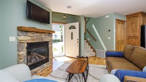 A living room with a stone fireplace, a television mounted above it, a wooden bench, and a sofa with blue cushions. - Downtown Fish Creek, Sleeps 6 - Yellow Birch Cottage (Fish Creek)