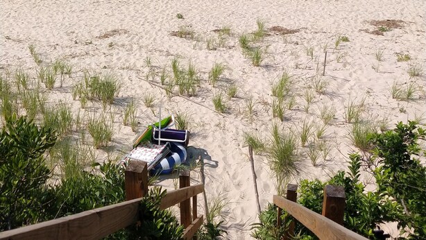 A sandy beach with a sailboat in the distance, a small inflatable boat, and a wooden fence leading to a beach chair with a towel.