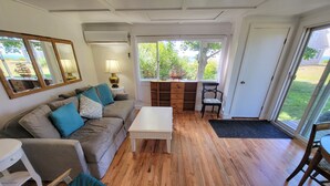 A living room with a grey sofa, a white coffee table, wooden flooring, a large window with a view of greenery, and a sliding glass door leading outside.