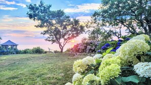 A gazebo in a grassy field with a tree and hydrangeas in the foreground.