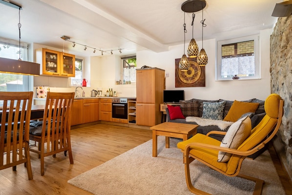 A modern kitchen with wooden cabinets, a dining area with a table and chairs, a sofa with yellow cushions, and a stone wall.