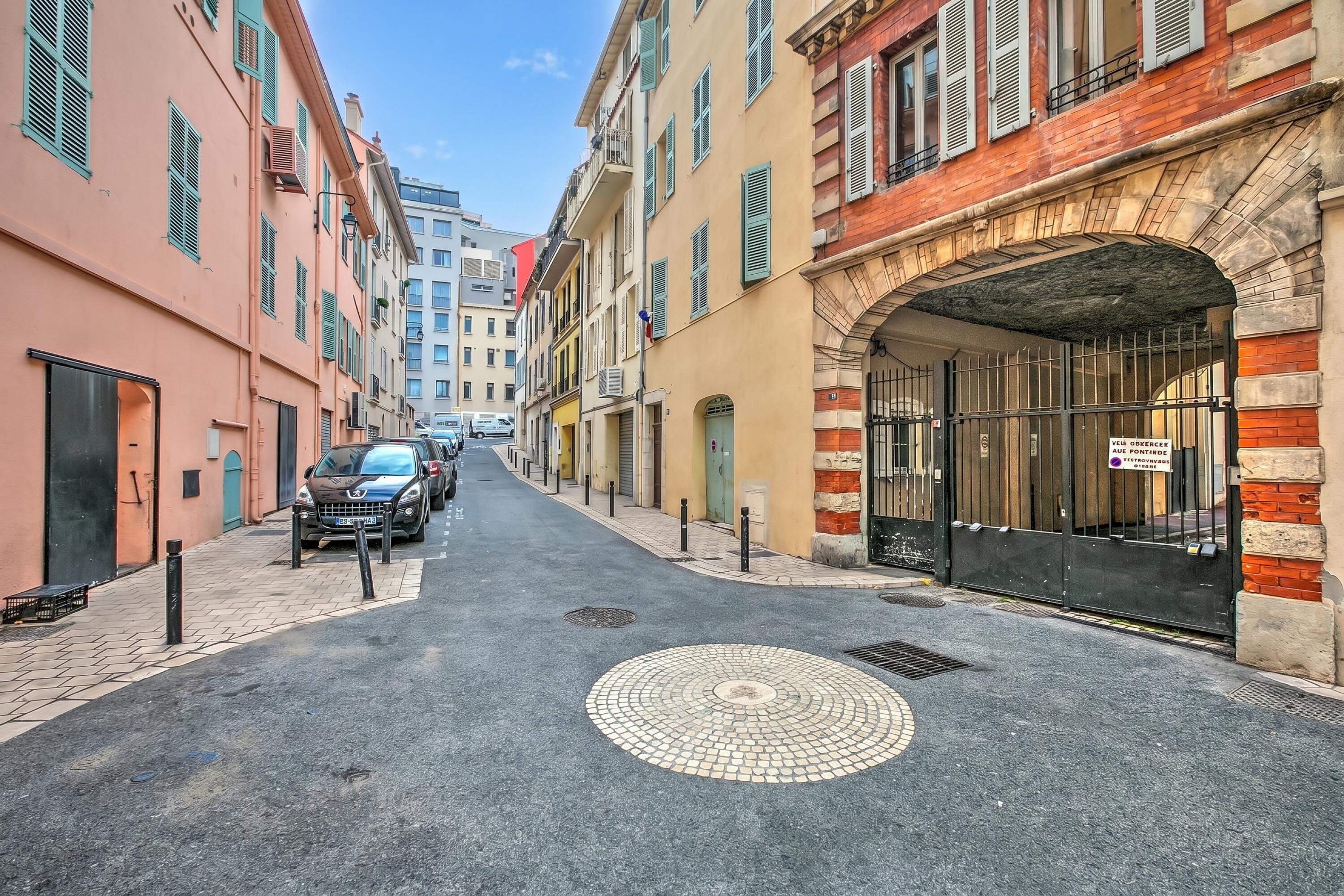 Une rue étroite bordée d’immeubles à plusieurs étages, une voiture garée et un regard d’égout.