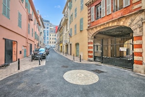 A narrow street with buildings on either side, a car parked on the left, and a black gate with a sign on the right.