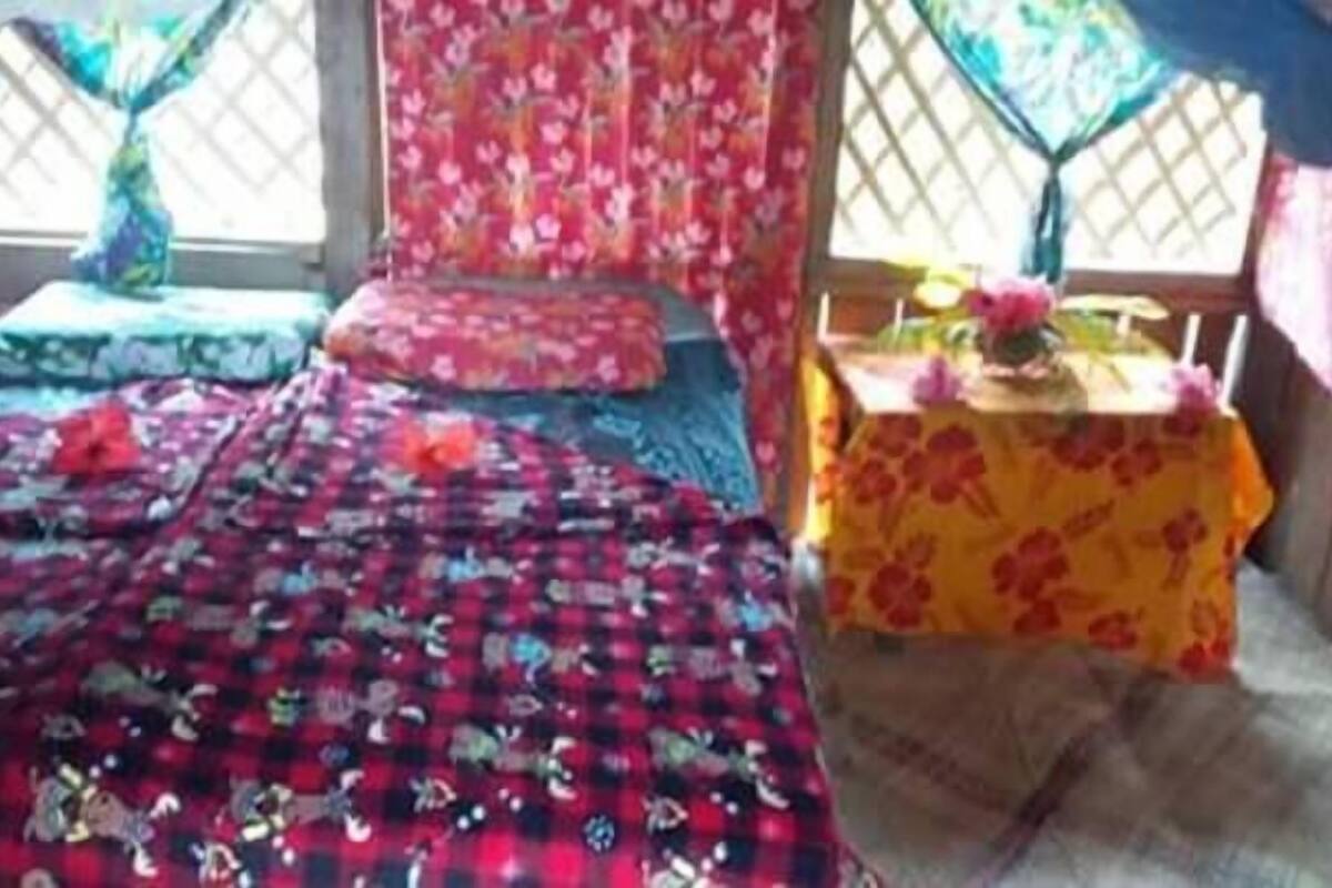 Une pièce comprenant un lit, une petite table recouverte d’une nappe à motifs floraux et une fenêtre ornée d’un croisillon.